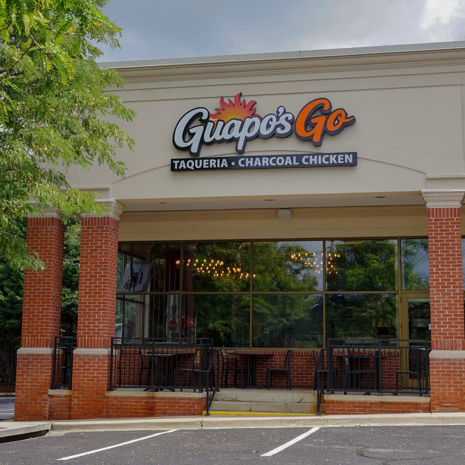 Guapo’s Go taqueria and charcoal chicken restaurant storefront with brick columns, outdoor seating, and tree-lined reflections.