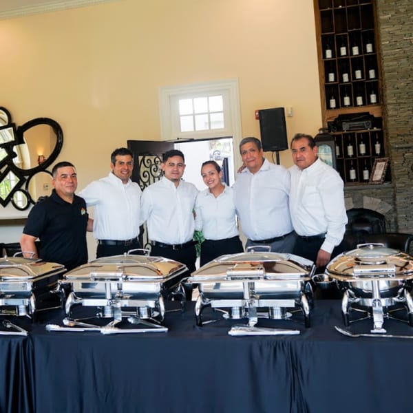 Guapo's GO catering staff, five men and one woman, standing behind chafing dishes on a black-draped table.