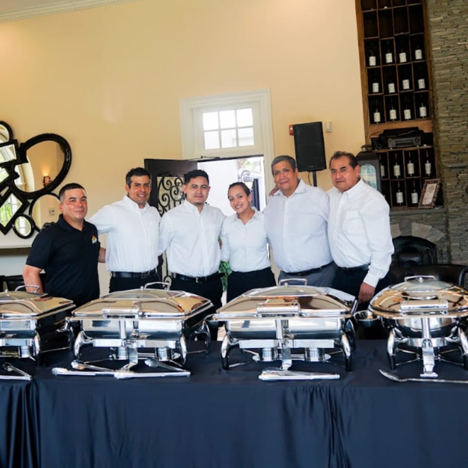 Guapo's GO catering staff, five men and one woman, standing behind chafing dishes on a black-draped table.