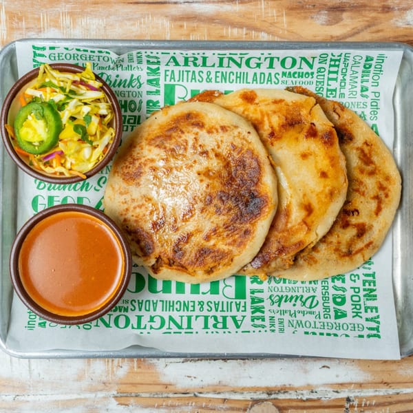 Three pupusas with a side of coleslaw and red sauce on a metal tray with branded paper.