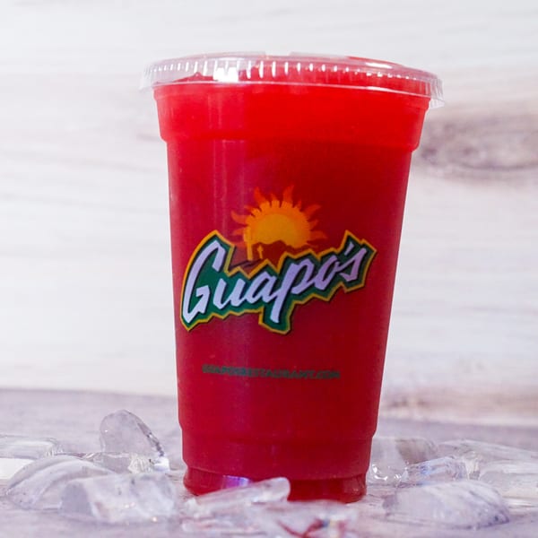A red Guapo's frozen drink in a clear plastic cup with ice cubes scattered around it.