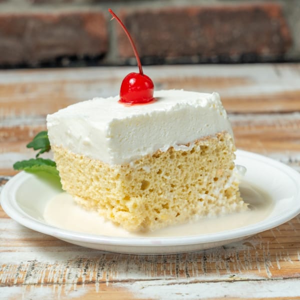 A slice of Tres Leches cake with whipped cream and a cherry on top, on a white plate.