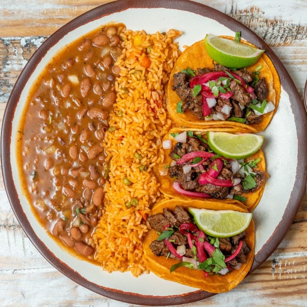 Three steak tacos with pickled onions, cilantro, and lime, served with rice and beans.