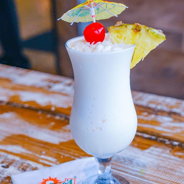 A frozen piña colada with a cherry, pineapple slice, and paper umbrella on a wooden table.