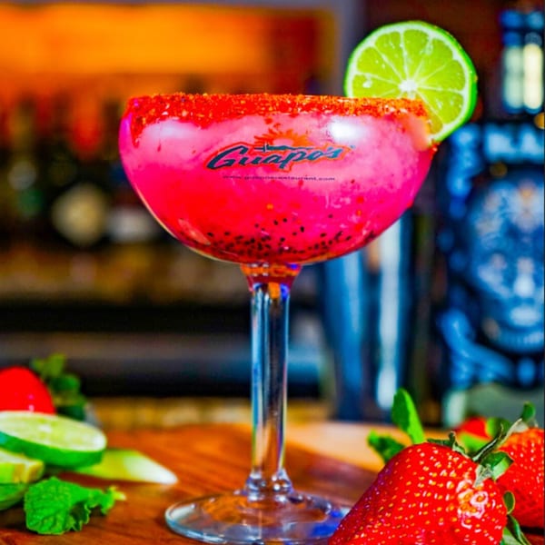 A Guapo's pink margarita with a chili-lime rim and a lime wedge, next to fresh strawberries.
