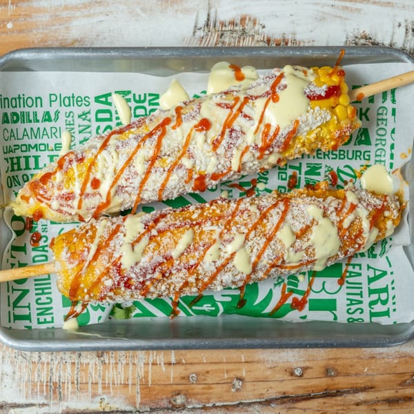 Two elotes, Mexican street corn, on a tray with white and orange sauce, and cotija cheese.
