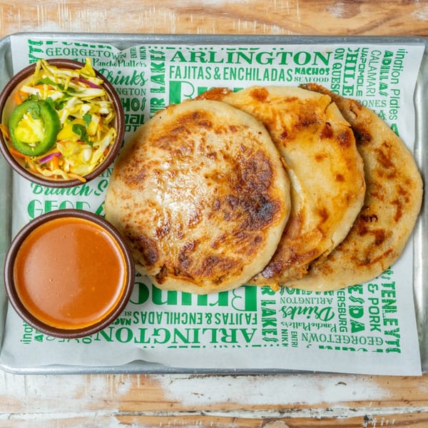 Three golden-brown pupusas with a side of slaw and red sauce on a branded tray liner.
