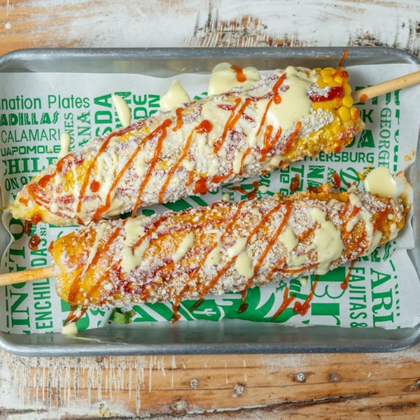 Two elotes, or Mexican street corn, on a tray with white and orange sauces and cotija cheese.