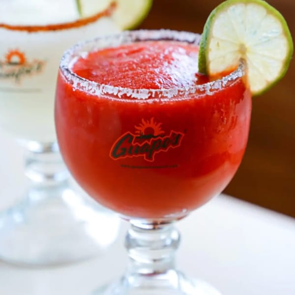 A Guapo's strawberry frozen margarita with a salted rim and lime wedge, with another drink blurred behind.